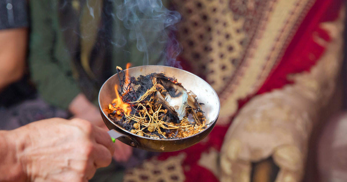 Peganum harmala (espand, uzerlik) seeds burning in a metal ladle, with rising smoke against a red Persian textile — the traditional ritual of burning wild rue to ward off the evil eye, practised across Iran and Central Asia for thousands of years.
